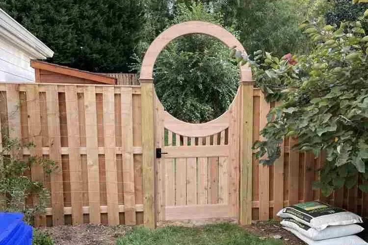 Wooden gate with circular archway set in a fence, surrounded by greenery.