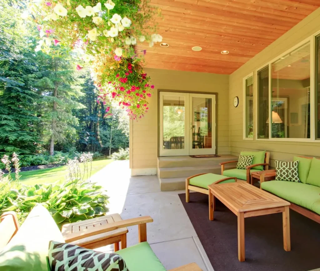 Covered patio with green cushioned wooden furniture and hanging flowers overlooking a lush garden.