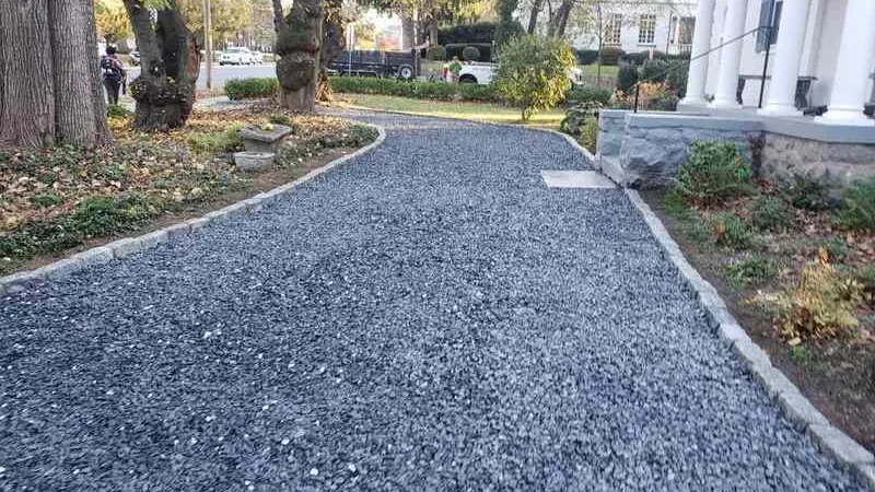 Gravel driveway leading to a columned entrance with trees and shrubs on the sides.