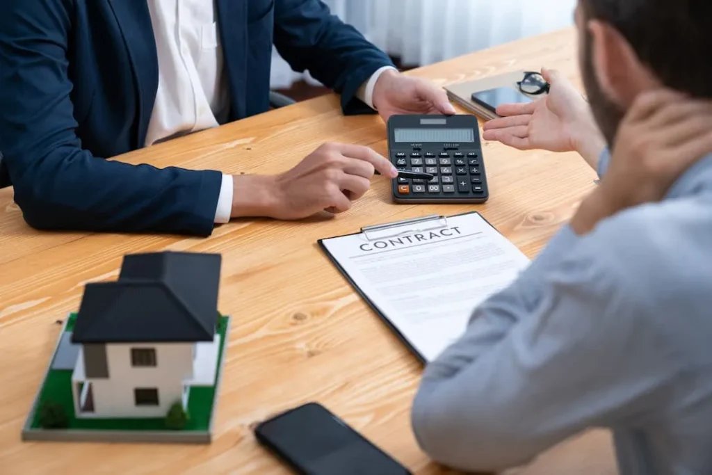 Two people discussing a contract with a calculator and house model on the table.