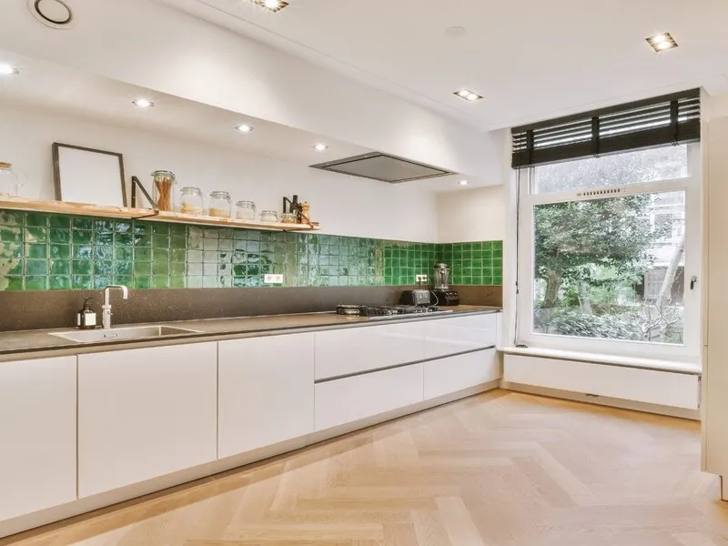 Modern kitchen with white cabinets, green tiled backsplash, and large window overlooking a garden.