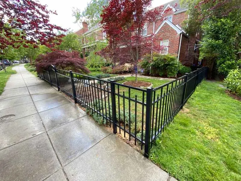 Types of Fences: Brick house with a black metal fence, trees, and a garden along a wet sidewalk.