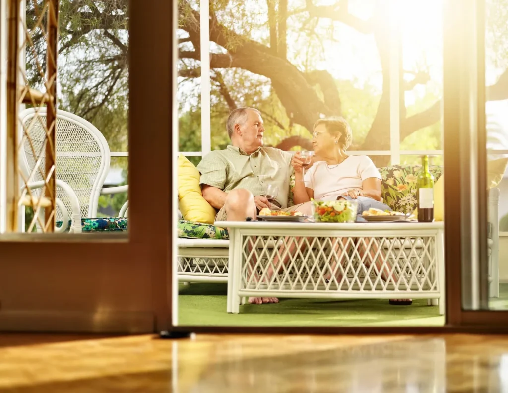 An elderly couple enjoys wine and conversation on a sunlit screened in patio.