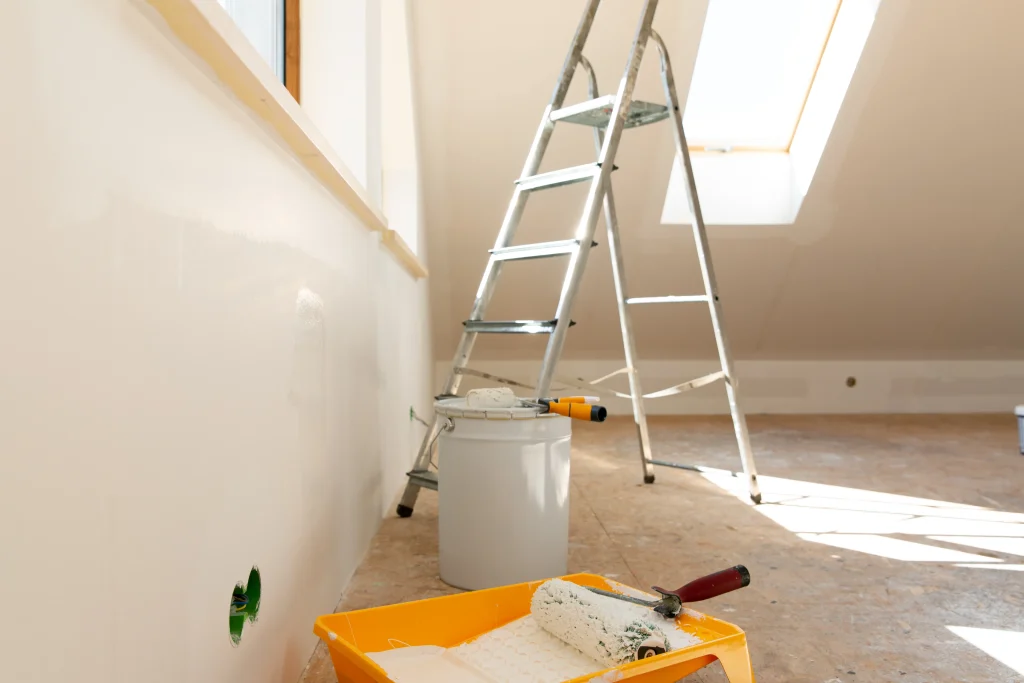 A room under renovation with a ladder, paint tray, and rollers near a skylight.