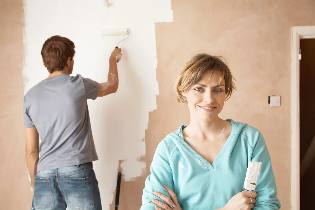 A woman smiles holding a paintbrush while a man paints a wall white.