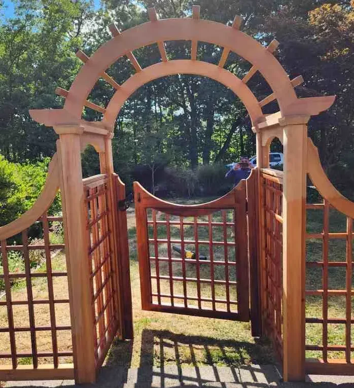 A wooden garden archway with an open gate under a sunny sky.