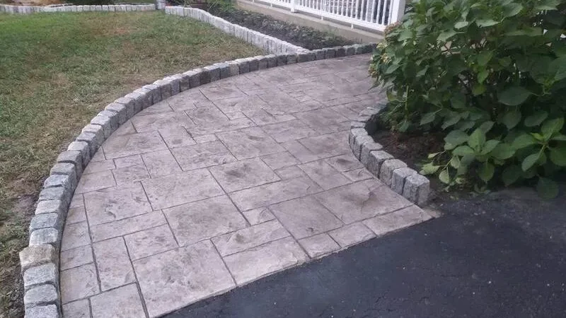 A curved, stamped concrete walkway bordered by bricks leads to a house with a porch.