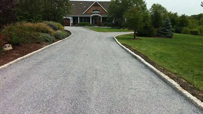 A tar and chip driveway leads to a brick house surrounded by trees and gardens.