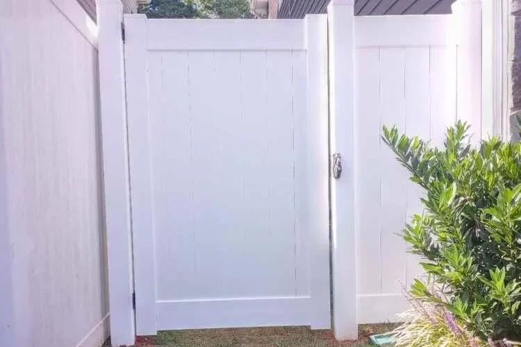 White gate with a latch between two fences, with grass and bushes in the foreground.