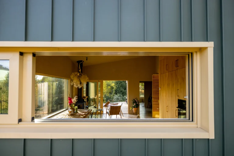 View through a window into a modern, well-lit room with a dining table and greenery outside.