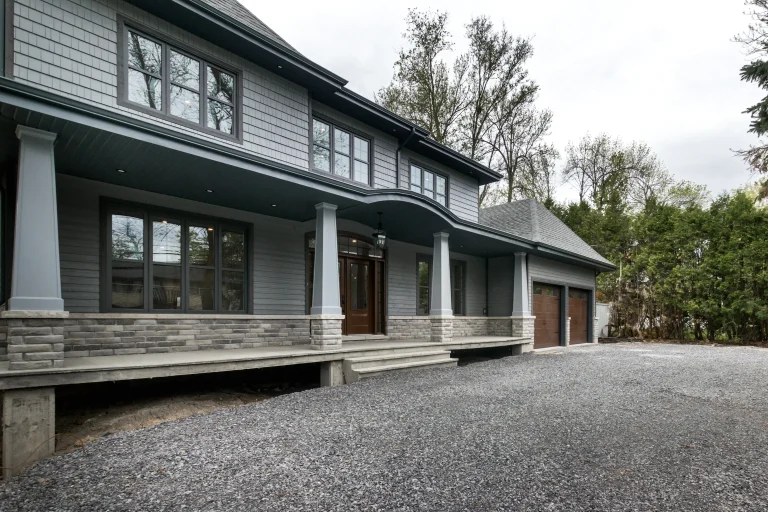 Gray two-story home addition with a covered porch and double garage, surrounded by trees.