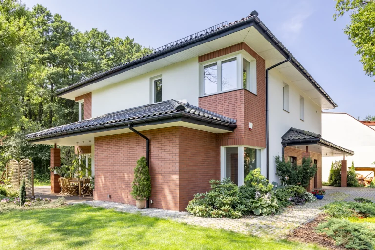 Modern two-story home addition with brick and white exterior, surrounded by lush greenery and a neatly maintained lawn.