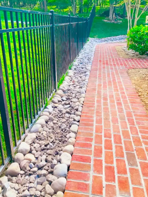 Red brick walkway by hardscape contractors featuring a river rock border and black metal fence in a Washington DC backyard