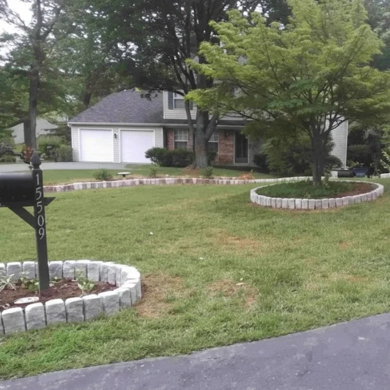 A front yard landscape featuring gray Belgian block stone edging for a mailbox planter, a circular tree ring, and a long curved garden border along the driveway.