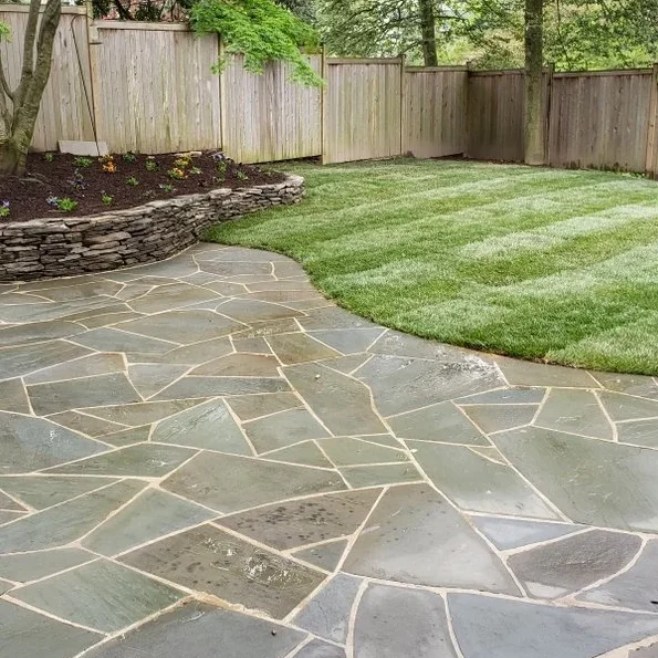 A high-end irregular flagstone patio transitioning into a lush lawn, featuring a hand-stacked natural stone retaining wall.