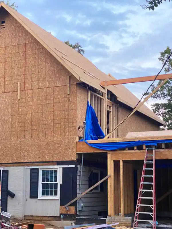 Home addition under construction with exposed wood framing and OSB sheathing, sloped roof in progress, ladder at the front, and blue tarp covering part of the new entry structure.