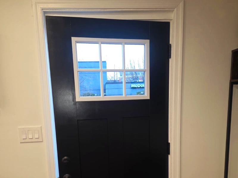 A newly remodeled entryway featuring a closed Craftsman matte black front door with a six-lite transom window at the top, matte black hardware, and new light wood plank flooring replacing the original carpet and doormat.