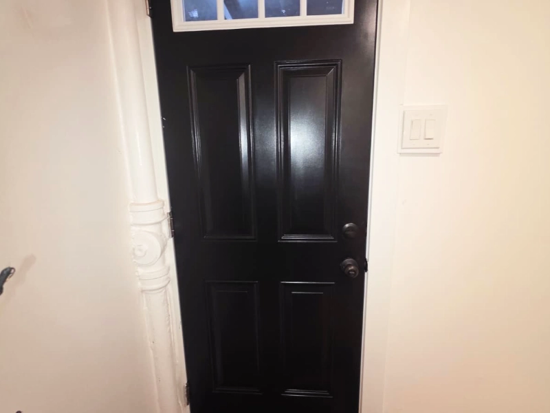 A newly remodeled interior entryway featuring a modern matte black four-panel door with a top transom window, crisp white trim, and a ribbed border coir doormat.