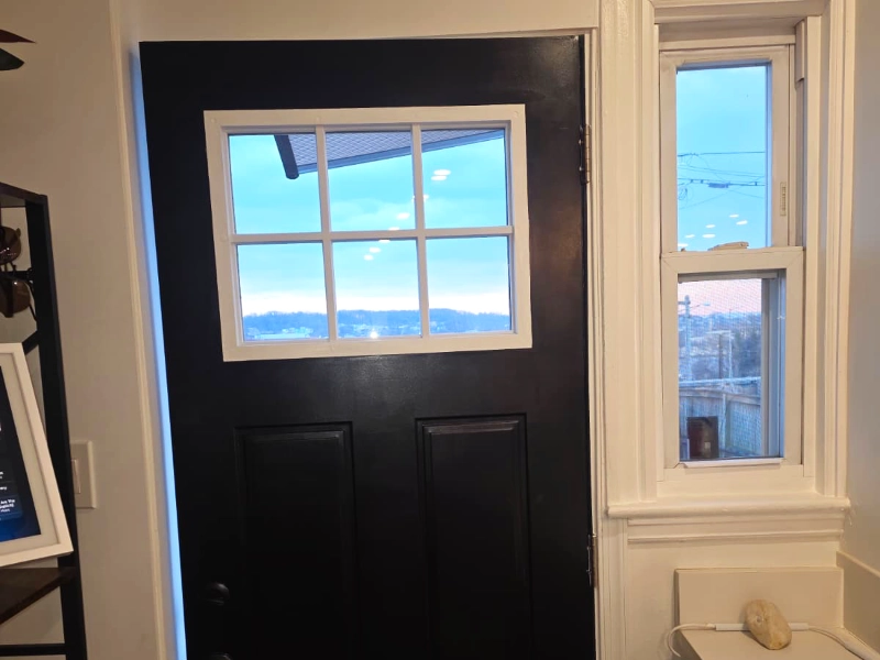 A newly installed matte black entry door featuring a top six-lite window grid, situated next to a crisp white-trimmed accent window and a light quartz countertop.