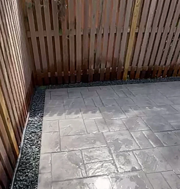 Finished stamped concrete patio installed beneath a raised rear porch with improved drainage, relocated access step, and underground downspout routing.
