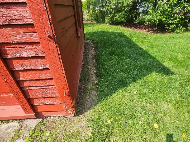 Side of a historic red barn before drainage work begins, showing grass and soil directly against the foundation where roof runoff was causing erosion concerns.