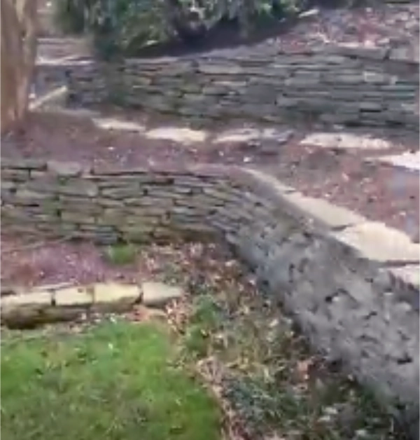 An angled view of sloped garden tiers with weathered stone walls before a cleanup, showing faded soil, scattered autumn leaves, and debris.