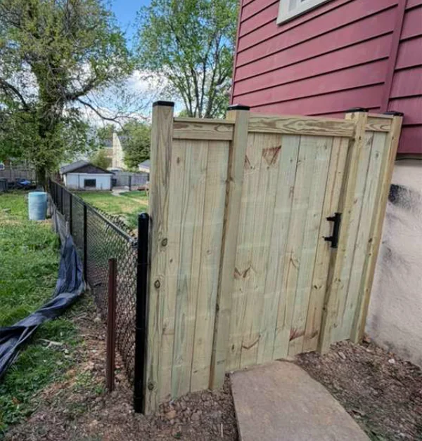 A newly installed four-foot black vinyl-coated chain-link fence connects directly to a wide six-foot pressure-treated wood gate next to a concrete walkway and red home siding.