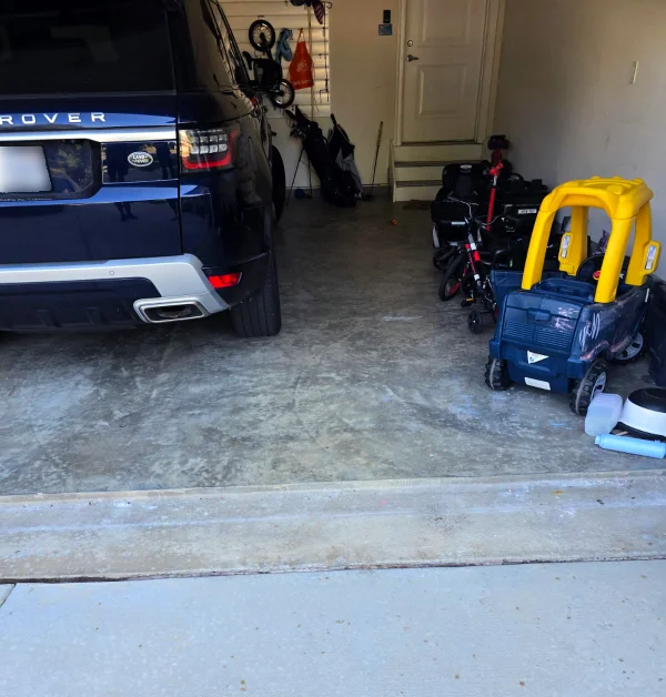 A cluttered residential garage with an unfinished, stained concrete floor features a parked dark blue SUV alongside scattered children's ride-on toys, bicycles, and golf clubs.