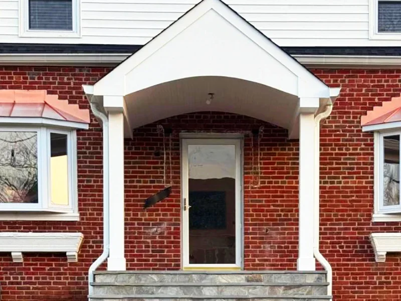A close-up of a completed white gabled entry portico with an arched underside, thick square columns, a gray flagstone landing, and adjacent white bay windows topped with copper roofs.