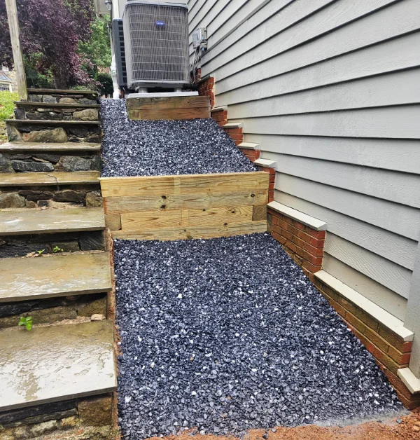 A completed hillside stabilization project features new timber retaining walls and dark gravel alongside stone steps leading to an HVAC unit.