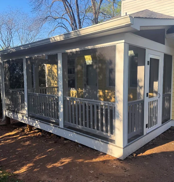 An exterior wide-angle shot of a newly built white screened-in porch extension featuring integrated roof skylights, large mesh screen panels, gray balustrade railings, and a pitched roof attached to a yellow home.