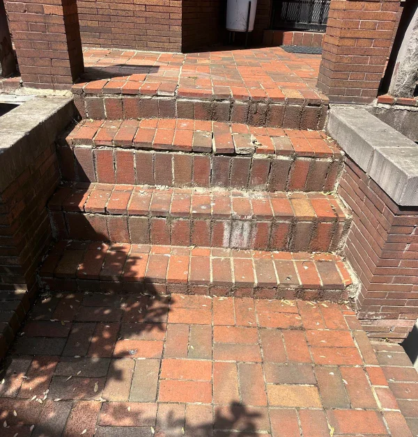 A high-angle view looking down at weathered red brick steps leading to a matching brick paver walkway dappled with afternoon tree shadows.