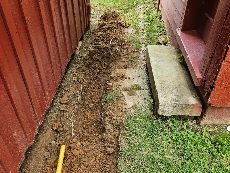 Close-up of a freshly excavated drainage trench alongside a historic barn foundation, prepared for installation of perforated pipe, filter fabric, and river gravel.