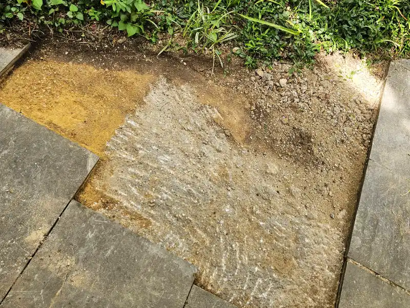 A top-down view of a section of a flagstone walkway cleared out to expose the leveled dirt and gravel base layer next to a green garden bed during a masonry repair project.