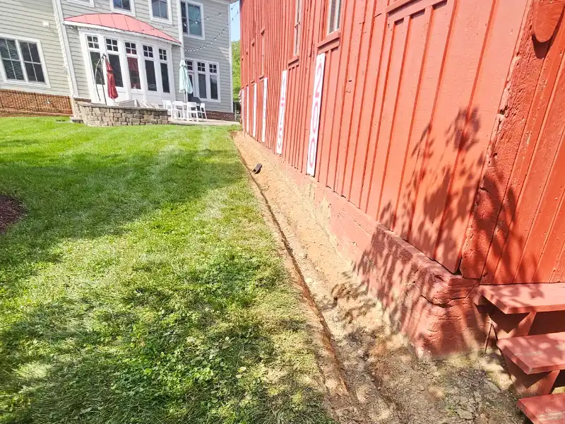 Freshly excavated trench running alongside a historic barn foundation before installation of drainage pipe and stone backfill.