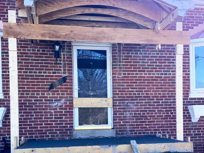 A construction photo showing a freshly poured concrete porch landing inside wooden forms below the rough timber framing for a new arched portico ceiling.