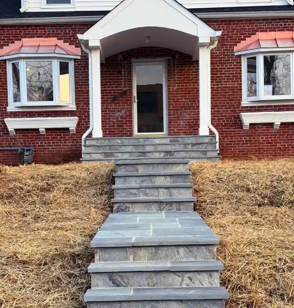 A renovated red brick home featuring a grand white gabled portico with square columns, a multi-tiered gray flagstone walkway leading down a straw-covered slope, and new white-trimmed bay windows with copper roofs.