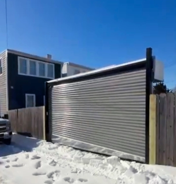 A heavy-duty industrial roll-up garage door made of galvanized steel with a black metal frame is seamlessly integrated into a wooden privacy fence along a snow-covered alley.