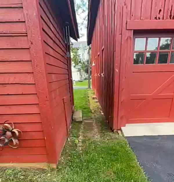 Before drainage improvements, grass and soil extend directly to the base of a historic barn with no gravel swale or drainage system present to manage roof runoff.