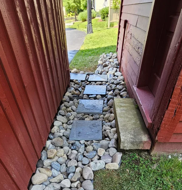 Completed river gravel drainage swale installed alongside a historic red barn, designed to capture roof runoff and direct water away from the foundation without using gutters.