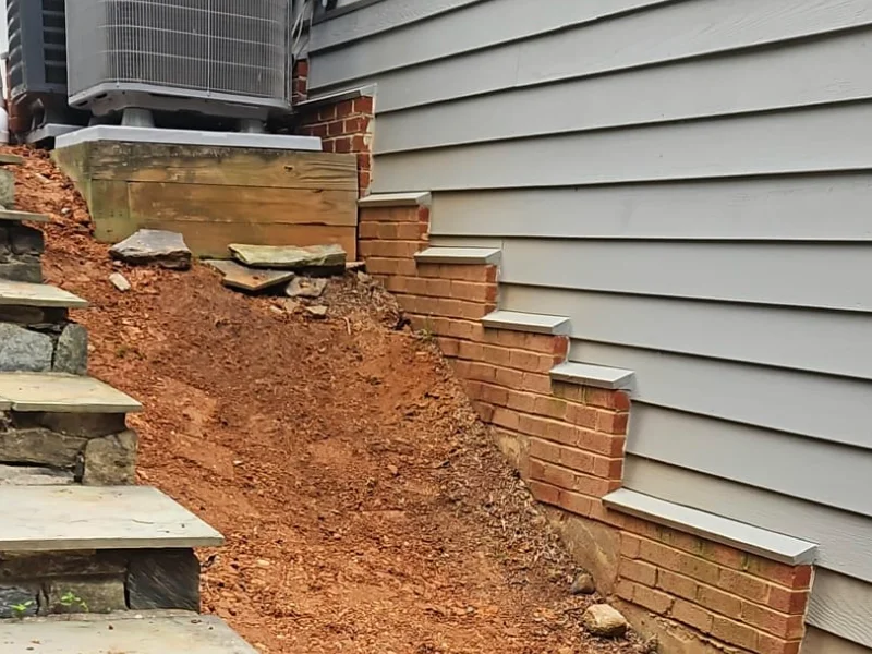 A worker uses a shovel to regrade and level the steep dirt slope next to outdoor stone steps during a stabilization project.