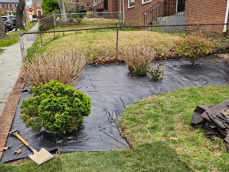 A shovel rests on freshly installed weed barrier fabric in a front yard garden bed during a comprehensive property cleanup and landscape reset.