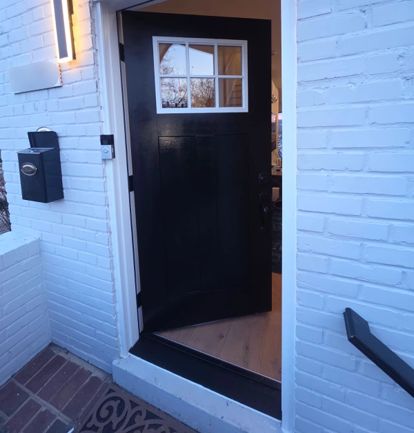 A slightly modern black craftsman-style front door with a six-pane window insert is set against a painted white brick wall featuring a vertical light fixture, a black mailbox, and a ring camera.