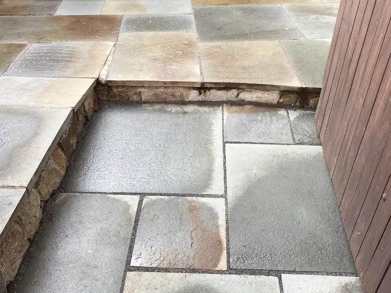 A section of a repaired flagstone patio showcases newly filled and damp, freshly activated polymeric sand joints running cleanly between level stone pavers next to a dark wood gate.