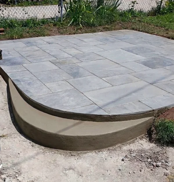 A full, angled view showcases a newly installed backyard patio made of rectangular, multi-sized gray flagstones, bordered by a freshly molded, smooth, and curved concrete step.