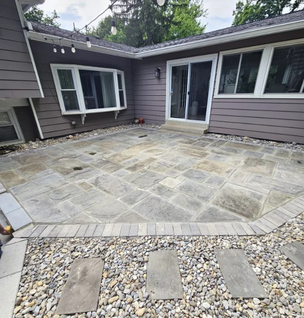 A newly restored Pennsylvania bluestone patio with freshly filled mortar joints sits behind a house with a sliding door, bordered by a river gravel swale featuring large rectangular stepping stones.