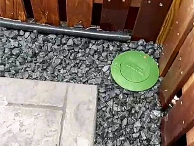 Green pop-up drain installed in the corner of the yard where the stamped concrete patio meets a black gravel drainage swale and wooden privacy fence.