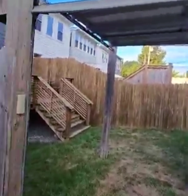 A weathered wooden support post and corrugated metal awning frame the view of a grassy backyard featuring a tall wooden fence and outdoor steps.
