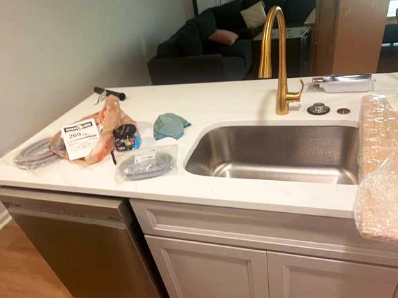 Kitchen island with a white quartz countertop, undermount sink, built-in dishwasher, and brushed gold faucet in the completed basement kitchen.