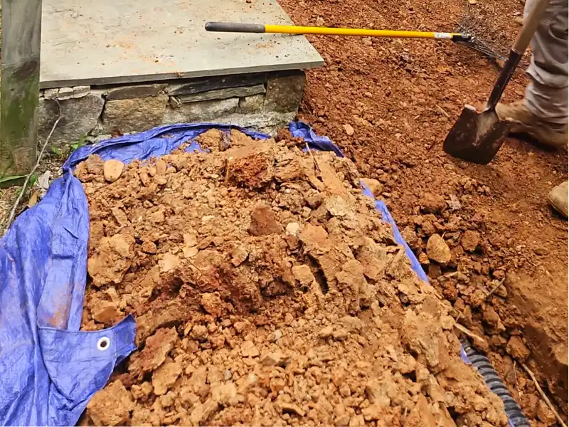 Landscaping tools rest on a freshly graded dirt slope where a drainage trench is being excavated alongside the home's foundation.